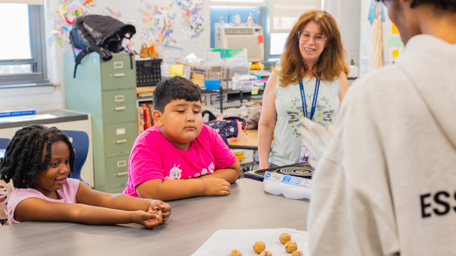 A smiling adult mentor and an older student watching over young children seated at a table with a tray of cookie dough