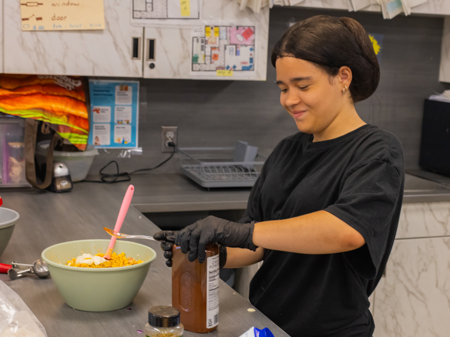A smiling student in black gloves spooning ingredients into a green mixing bowl of street corn
